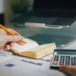 Freelancer pracuje u notebooku na faktury do zahraničí s papíry, poznámkami a kalkulačkou